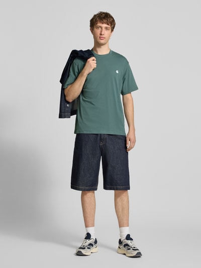 Męski model nosi T-shirt z wyhaftowanym logo model 'Madison' od Carhartt Work In Progress z bawełny, krój regular fit, okrągły dekolt, długość tyłu w rozmiarze M: 75 cm, pełen widok z przodu.