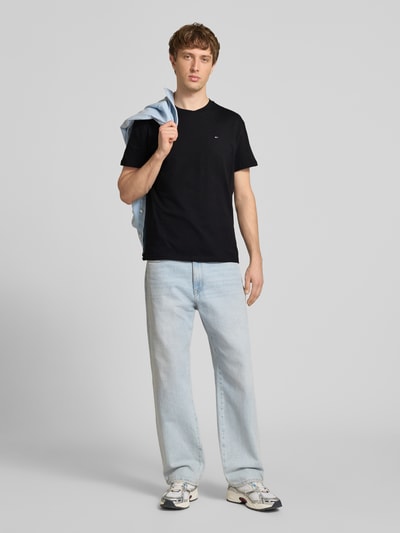 Männermodel trägt Regular Fit T-Shirt aus reiner Baumwolle von Tommy Jeans in Schwarz, unifarben, Logo-Stitching, Ganzkörperansicht von vorne.