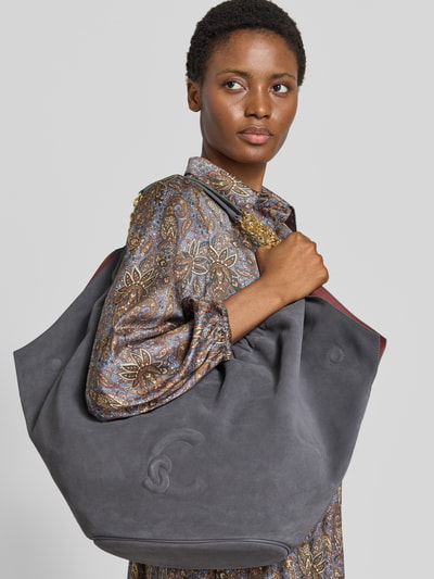 Frauenmodel trägt Handtasche mit Logo-Detail Modell 'Smash' von Coccinelle in Grau mit Ketten-Detail, Nahaufnahme Oberkörper, Magnetverschluss, KI-modifiziertes Bild.
