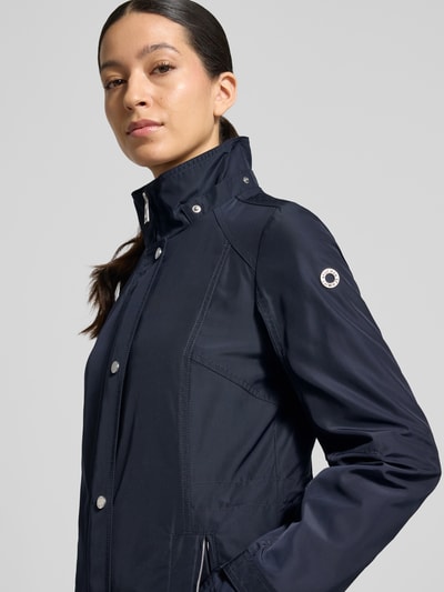Frauenmodel trägt Regular Fit Jacke mit Logo-Print von Fuchs Schmitt in Dunkelblau, glatte Textur, sichtbarer Stehkragen, Nahaufnahme Oberkörper. Maße und Details laut Größe 36: 71 cm Rückenlänge, 40 cm Schulterbreite, 51 cm Brustweite, 66 cm Ärmellänge, Kapuze mit Tunnelzug, Reißverschlusstaschen.