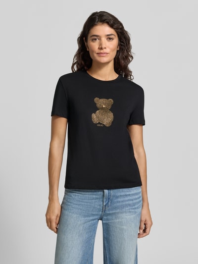 Kobiece model nosi Slim Fit T-Shirt mit Ziersteinbesatz od Liu Jo White z motywem z ozdobnymi kamieniami oraz okrągłym dekoltem, czarna bawełna, długość tyłu w rozmiarze S 57 cm, Ganzkörperansicht von vorne.