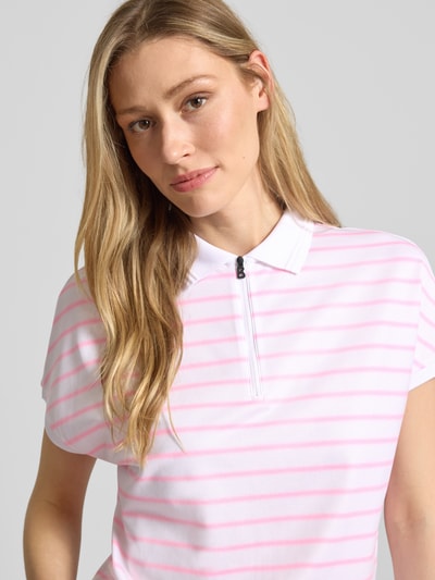 Modelka nosi Regular Fit Poloshirt mit Reißverschluss von FIRE + ICE, wzór na całej powierzchni, tunel, długość tyłu w rozmiarze S 62 cm, zamek błyskawiczny, model 'GAJA2', różowe paski na białym tle, widok z przodu, zbliżenie na górną część ciała.
