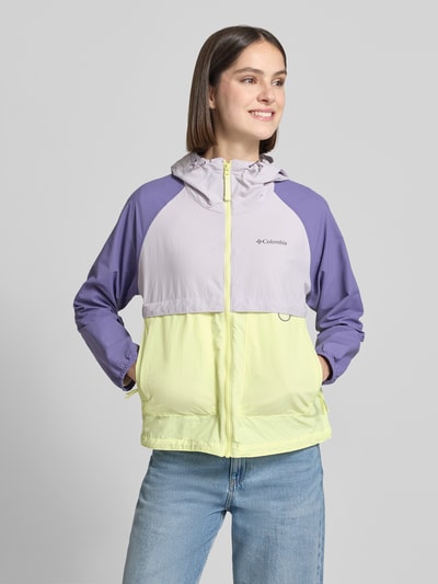 Frauenmodel trägt Windbreaker mit Rückenbelüftungssystem und Kapuze von Columbia in Flieder, Gelb und Lila, Lockerer-Schnitt, Ganzkörperansicht von vorne. Omni-Shield™ Technologie, Schmutzabweisend, Kinnschutz, Eingrifftaschen mit Reißverschluss, elastische Bündchen, Saum mit Kordelzug, Logo-Print, Rückenlänge 59 cm, Schulterbreite 45 cm, Brustweite 51 cm, Ärmellänge 57 cm.