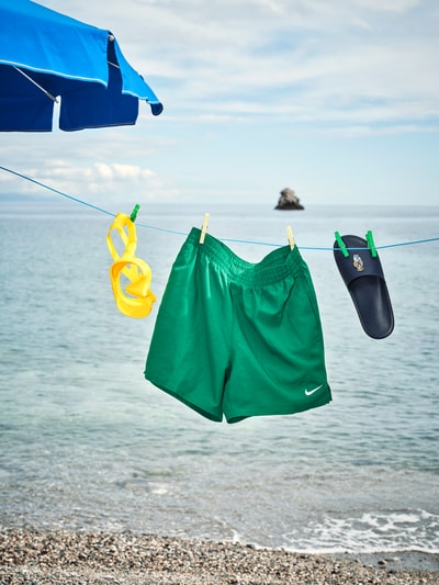 Produktfoto, grüne Badeshorts mit Logo-Stitching und Eingrifftaschen von Nike, Relaxed-Fit, elastischer Bund, am Strand, steiniger Untergrund, blauer Himmel, ruhiges Meer, Schnorchel und Sandale als Accessoires