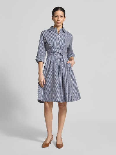 Modelka nosi Hemdblusenkleid mit Taillenband od Apricot, bawełna z elastanem, krój slim fit, długość midi, kołnierz kent, pasek, rękaw 3/4, długość tyłu w rozmiarze S: 110 cm, szerokość w rozmiarze S: 40 cm, długość rękawa w rozmiarze S: 50 cm, wzór w paski, ciemnoniebieskie i białe odcienie, gładka tekstura, pełen widok z przodu.