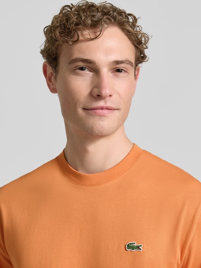 Mannenmodel draagt Regular Fit Poloshirt aus reiner Baumwolle van Lacoste in effen oranje met logo-stitching, rondhals, korte mouw, materiaal katoen. Nahaufnahme bovenlichaam.