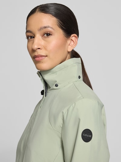 Frauenmodel trägt Parka mit Kapuze und verdecktem Reißverschluss von G.I.G.A. DX in hellem Grün, Label-Patch am Ärmel, Nahaufnahme Oberkörper. Wassersäule 10.000mm, gerader Schnitt, Kordelstopper, seitliche Reißverschlusstaschen, Rückenlänge 70 cm, Schulterbreite 41 cm, Brustweite 51 cm, Ärmellänge 63 cm.