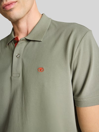 Mężczyznamodel nosi Regular Fit Poloshirt aus reiner Baumwolle von Tom Tailor, oliwkowy kolor, krój regular fit, pikowana bawełna, logo na piersi, dwuguzikowe zapięcie, polokołnierzyk, krótki rękaw, widok z przodu, zbliżenie na górną część ciała.