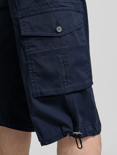 Męski model nosi Capri Cargo Pants mit Pattentaschen od bugatti, granatowy bawełniany materiał, prosty krój straight fit, duże kieszenie, detal na nogawce, zbliżenie na nogawkę