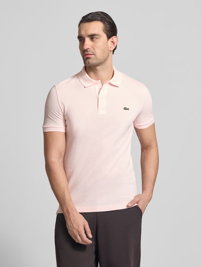 Männermodel trägt Slim Fit Poloshirt aus reiner Baumwolle von Lacoste, efekt: jednolity kolor, krój slim fit, wyhaftowane logo, kołnierzyk polo, krótki rękaw, prążkowane zakończenie rękawów, materiał: pika z bawełny, jasnoróżowa barwa, gładka faktura, całkowity widok z przodu