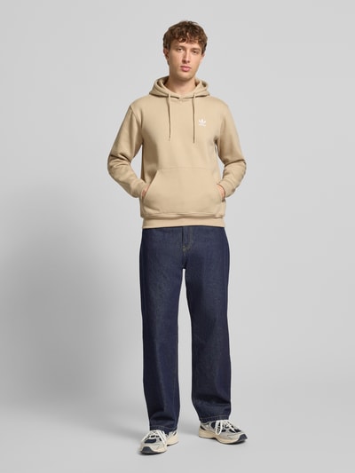 Mężczyznamodel nosi Hoodie mit Känguru-Tasche od adidas Originals, mieszanka bawełny, krój regular fit, haftowane logo, kieszeń kangurka, całkowity widok z przodu.