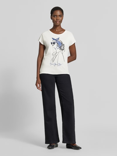 Frauenmodel trägt T-Shirt aus Baumwoll-Viskose-Mix von Christian Berg Woman in Offwhite mit schwarzem Grafikdruck, schwarze Hose, Ganzkörperansicht von vorne. Gerader Schnitt, Rundhalsausschnitt, überschnittene Schultern, Rückenlänge bei Größe S 56 cm.