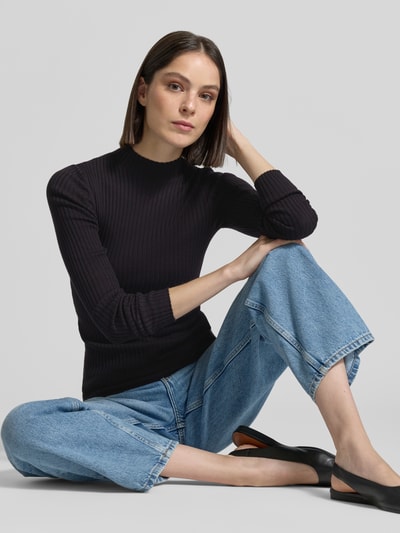 Vrouwenmodel draagt gebreide pullover uit puur katoen model 'ALAANIA' van Armedangels, look: effen, slim fit, labelpatch, logostitching, mock neck, hooggesloten hals, lange mouw, materiaalverwerking: knitwear, kleur: zwart, voorkantperspectief, halve lichaamsopname.