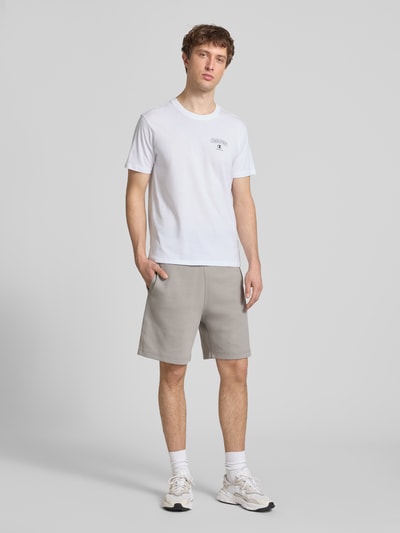 Männermodel trägt T-Shirt mit Logo und Rundhalsausschnitt von CHAMPION in Weiß, reine Baumwolle, Regular Fit, Ganzkörperansicht von vorne.