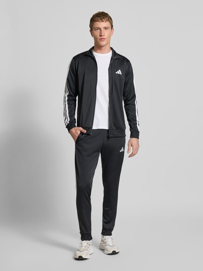 Męski model nosi Trainingsanzug mit Stretch-Anteil od adidas Sportswear, czarny z białymi detalami, elastyczna mieszanka materiałów, prosty krój, stójka, zamek błyskawiczny, elastyczne wykończenia oraz pas, wpuszczane kieszenie, detale z logo; szerokość w rozmiarze M: 39 cm, długość tyłu: 70 cm, szerokość ramion: 44 cm, długość rękawów: 65 cm, długość wewnętrzna nogawki: 73 cm; całopostaciowy widok z przodu.