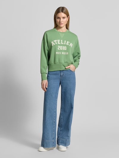 Kobieta modelka nosi Relaxed Fit Sweatshirt aus Viskose-Mix Modell 'Nima' od MOS MOSH, elastyczna mieszanka wiskozy, krój relaxed fit, detal z logo, okrągły dekolt, elastyczne wykończenia, zielony kolor, gładka faktura, widok całej sylwetki z przodu.