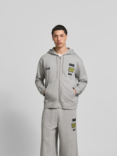 Männermodel trägt Regular Fit Sweatjacke mit Logo-Patch von MM6 Maison Margiela in Grau, reine Baumwolle, Ganzkörperansicht von vorne, Herren Sweatjacke mit Kapuze, Tunnelzug und Reißverschluss, Rückenlänge 69 cm, Schulterbreite 45 cm, Ärmellänge 72 cm.