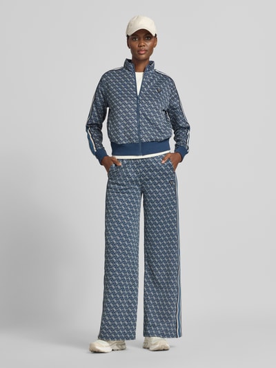 Frauenmodel trägt Sweatpants mit Logo-Muster im Allover-Look von GUESS Activewear in Blau und Weiß, Wide Leg, elastischer Bund, Material-Mix, Ganzkörperansicht von vorne.