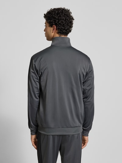 Männermodel trägt Slim Fit Trainingsanzug mit Logo-Stitching von adidas Sportswear in Dunkelgrau, glatte Textur, Ganzkörperansicht Rückansicht, Stehkragen, Eingrifftaschen, Reißverschluss, elastische Abschlüsse, Bundweite 39 cm, Rückenlänge 71 cm, Schulterbreite 47 cm, Ärmellänge 66 cm, Innenbeinlänge 76 cm.