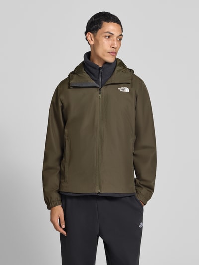 Męski model nosi kurtkę Regular Fit Jacke mit Logo-Print Modell 'Quest' od The North Face w odcieniu oliwkowym, z kapturem ze stójką, wpuszczanymi kieszeniami oraz zamkiem błyskawicznym, długość tyłu w rozmiarze M: 72 cm, szerokość ramion w rozmiarze M: 46 cm, szerokość klatki piersiowej w rozmiarze M: 60 cm, długość rękawów w rozmiarze M: 69 cm, ujęcie całej sylwetki z przodu.
