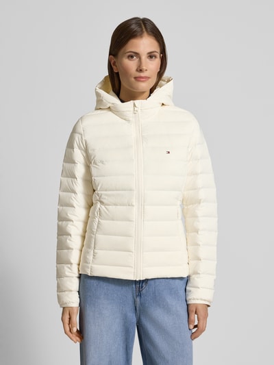 Kobiece modelka nosi Kurtka puchowa Slim Fit Daunenjacke mit abnehmbaren Kapuze od Tommy Hilfiger, jednolity kolor, logo, stójka, kaptur, długi rękaw, elastyczne zakończenia rękawów, hydofobowy materiał, kieszenie zapinane na zamek błyskawiczny; pełny widok od przodu.
