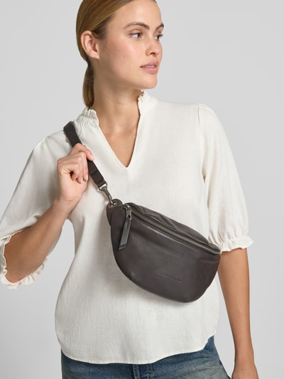Frauenmodel trägt Bauchtasche mit Logo-Prägung Modell 'Tavia' von LIEBESKIND BERLIN, abnehmbarer Schulterriemen, Hauptfach mit Reißverschluss, Farbe Dunkelbraun, glatte Textur, Breite 30 cm, Länge 15 cm, Tiefe 2 cm, Länge Schulterriemen 87 cm, Ganzkörperansicht von vorne.