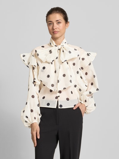 Vrouwenmodel draagt Regular Fit strikblouse met ruches model 'DOROTHY' van Vero Moda, all-over polkadots, semi-transparant, lange ballonmouwen, elastische mouwboord, mock neck met strik, eenrijige knoopsluiting, all-over motief, applicatie met ruches, regular fit pasvorm, voorkant volledige lichaamsaanzicht.