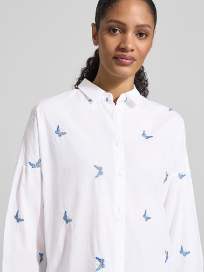 Modelka kobieca nosi Regular Fit Bluse mit Stitchings Modell 'LUCY' od 0039 Italy z czystej bawełny, biały materiał z niebieskimi haftami, kołnierz koszulowy, zapięcie na guziki, krój regular fit, długość tyłu w rozm. S: 66 cm, długość rękawa w rozm. S: 46 cm; zbliżenie na górną część ciała