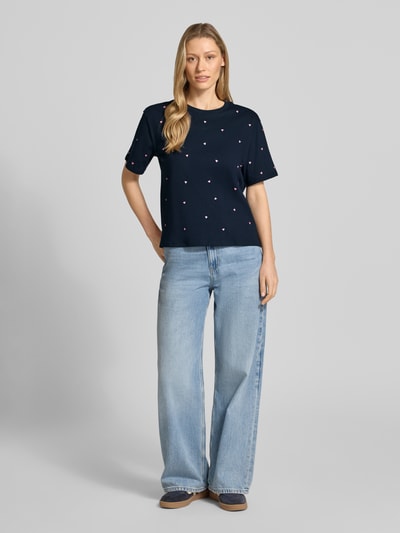 Frauenmodel trägt Boxy Fit T-Shirt aus reiner Baumwolle von Tom Tailor Denim, unifarben mit Allover-Herz-Stickerei, kurzarm, Rundhals, Seitenschlitze, kombiniert mit hellblauer Jeans; Ganzkörperansicht von vorne.