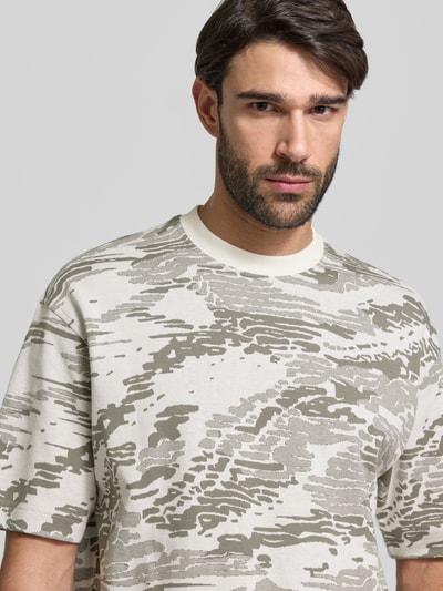 Männermodel trägt Regular Fit T-Shirt aus reiner Baumwolle Modell 'TE_WILDNATURE' von BOSS Orange mit abstraktem Allover-Muster in Beige und Grau, fein gestrickt, Rundhals, Label Patch, Nahaufnahme Oberkörper.
