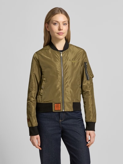 Frauenmodel trägt Bomberjacke mit Label-Patch und Reißverschlusstasche am Ärmel von Original Bombers in Oliv mit schwarzen elastischen Abschlüssen, Lockerer-Schnitt, Stehkragen, Reißverschluss und Logo-Stitching, seitliche Eingrifftaschen mit Druckknopfverschluss, Ganzkörperansicht von vorne.