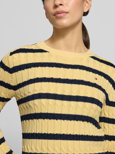 Frauenmodel trägt Slim Fit Strickpullover aus reiner Baumwolle von Tommy Hilfiger, gelb-blau gestreift, Logo-Detail, Nahaufnahme Oberkörper.