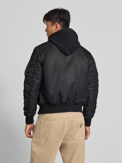 Männermodel trägt Jacke mit Reißverschlusstasche am Ärmel von Alpha Industries in Schwarz, glatte Textur, Bündchen gerippt, Ganzkörperansicht Rückansicht. Rückenlänge 64 cm, Schulterbreite 54 cm, Brustweite 54 cm, Ärmellänge 72 cm, Regular Fit, Kapuze, Label-Patch, Eingrifftaschen.