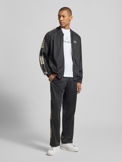 Männermodel trägt schwarze Trainingsjacke mit Zweiwege-Reißverschluss von EA7 Emporio Armani, Regular-Fit, Logo, Stehkragen, Eingrifftaschen mit Druckknopfverschluss, Rückenlänge 68 cm, Ärmellänge 57 cm, Ganzkörperansicht von vorne.
