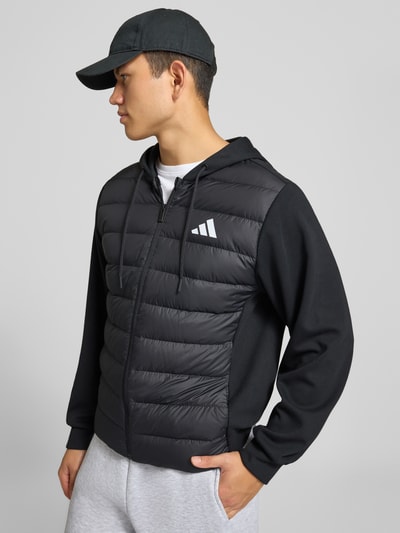 Männermodel trägt Regular Fit Steppjacke mit Kapuzenkragen von adidas Sportswear in Schwarz mit glatter und gesteppter Textur, Ganzkörperansicht Vorderansicht.