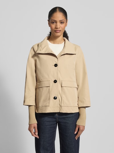 Frauenmodel trägt Jacke mit Stehkragen und Pattentaschen von Christian Berg Woman Selection, krój regular fit, beżowy kolor, duże czarne guziki, szerokie mankiety, dwie duże kieszenie z patkami, szerokość w rozmiarze 36: 46 cm, długość tyłu: 60 cm, szerokość ramion: 40 cm, długość rękawa: 67 cm, jeansy, prosty krój, gładka tekstura, całkowity widok z przodu