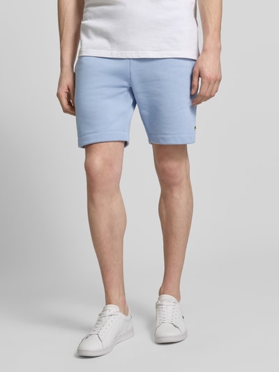 Mannenmodel draagt Regular Fit Sweatshorts aus Baumwoll-Mix van Lacoste in lichtblauw, un kleur, sweatstof, elastische tailleband, voorzakken, regular fit, label patch, detailopname benen, vooraanzicht.