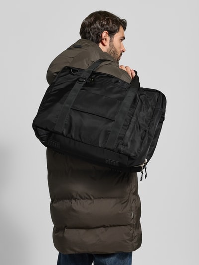 Männermodel trägt Tasche mit Logo-Detail von AY STUDIOS in Schwarz, große Netztaschen, rechteckige Form, Ganzkörperansicht Rückansicht. Hauptfach mit Reißverschluss, verstellbarer Schulterriemen, Maße: 48x35x22 cm, Länge Schulterriemen 100 cm.