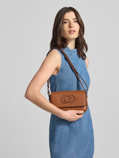 Kobiece model nosi torebkę w imitacji skóry model 'Isola' od Guess, kolor brązowy, detal z logo, główna przegroda z klapką, regulowane ramiączko, zapięcie na magnes, szerokość w rozmiarze 1: 23 cm, długość w rozmiarze 1: 16 cm, głębokość w rozmiarze 1: 6 cm, długość paska: 73 cm, zdjęcie całej sylwetki z przodu.