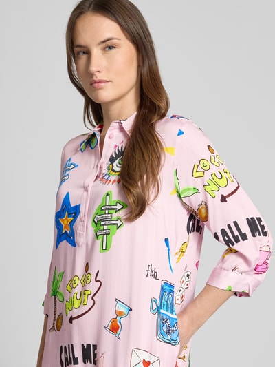 Frauenmodel trägt Relaxed Fit Hemdblusenkleid aus reiner Viskose von Milano Italy, rosa Grundfarbe, bunte Comic-Motive, Knopfleiste, 3/4-Arm, Nahaufnahme Oberkörper.