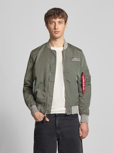 Männermodel trägt Regular Fit Bomberjacke mit Logo-Patch von Alpha Industries in Olivgrün mit Stehkragen und Reißverschlüssen, Nahaufnahme Oberkörper.