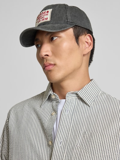 Męski model nosi Base Cap mit Logo-Patch od Closed, czarna czapka z daszkiem z bawełny, tkanina stylizowana na denim, wyhaftowany napis, regulowane zapięcie, naszywka z logo z przodu, widok z przodu z bliska.