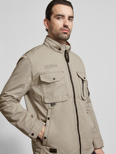 Männermodel trägt Jacke aus reiner Baumwolle mit Brusttaschen von camel active in beige mit Brusttaschen, recht pasvorm, opstaande kraag, kinbescherming, ritssluiting, borstzakken en steekzakken, mouwzak en mouwsluitingen, labelpatch; ruglengte maat 50: 73 cm, schouderbreedte: 52 cm, borstwijdte: 58 cm, mouwlengte: 64 cm. Nahaufnahme Oberkörper.