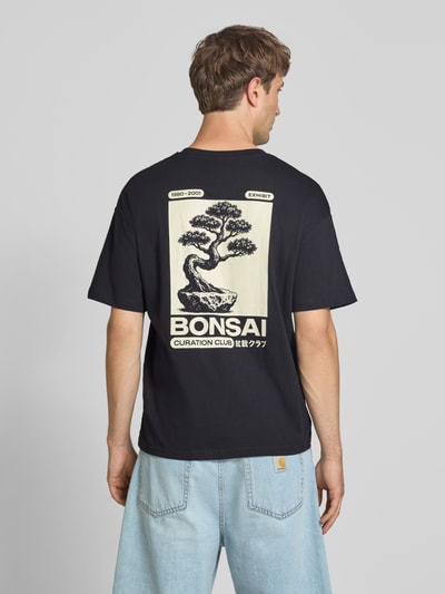 Männermodel trägt Relaxed Fit T-Shirt aus reiner Baumwolle Modell "DYLAN" von JACK & JONES, schwarzer Stoff mit Bonsai-Print, Ganzkörperansicht Rückansicht.