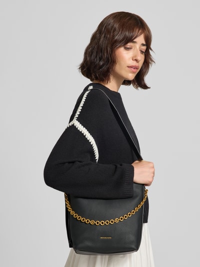 Frauenmodel trägt Handtasche aus echtem Leder Modell 'Nolita' von MICHAEL Michael Kors in Schwarz mit Ketten-Optik, Nahaufnahme Oberkörper. Hauptfach mit Magnetverschluss, verstellbarer Schulterriemen, Logo-Detail, Maße 28x25x10 cm, Riemenlänge 80 cm.