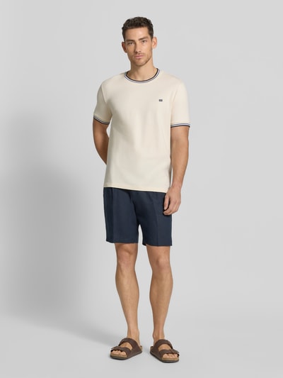 Męski model nosi T-Shirt mit Kontraststreifen und Rundhalsausschnitt von Christian Berg Men, bawełna z domieszką elastanu, krój regular fit, kontrastowe paski, okrągły dekolt, długość tyłu w rozmiarze M: 71 cm, jasny beż, granatowe szorty, brązowe sandały, widok całej sylwetki z przodu.