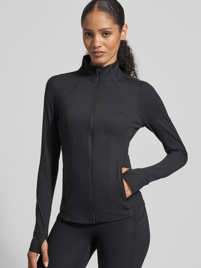 Frauenmodel trägt Slim Fit Jacke mit Stehkragen Modell 'Storm' von Fabletics im Elastischer-Material-Mix mit Daumenschlaufen, Kinnschutz, Schwarz, Nahaufnahme Oberkörper. Rückenlänge 61 cm, Schulterbreite 34 cm, Brustweite 43 cm, Ärmellänge 66 cm bei Größe S.