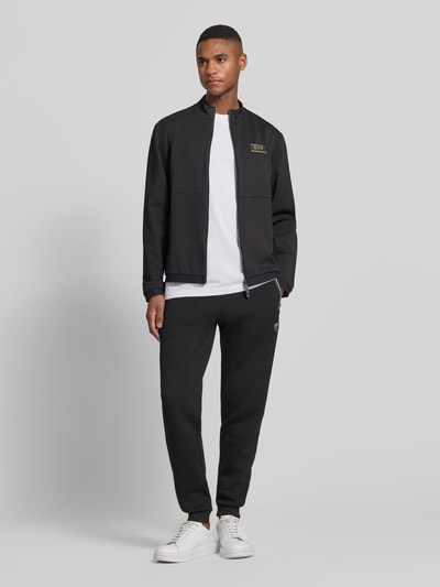Męski model nosi Bluza rozpinana z zamkiem błyskawicznym dwustronnym od EA7 Emporio Armani, mieszanka materiałów z dodatkiem wiskozy, krój regular fit, stójka, naszywka z logo, kieszenie zapinane na zamek błyskawiczny, długość tyłu 65 cm, długość rękawa 65 cm, czarna, gładka faktura, cała sylwetka z przodu.