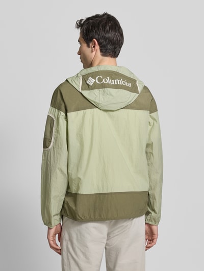 Männermodel trägt Windbreaker mit verdecktem Reißverschluss von Columbia in Grün mit Logo auf Kapuze, Regular-Fit, Rückansicht Ganzkörperansicht. Rückenlänge 70 cm, Schulterbreite 64 cm, Brustweite 57 cm, Ärmellänge 56 cm.