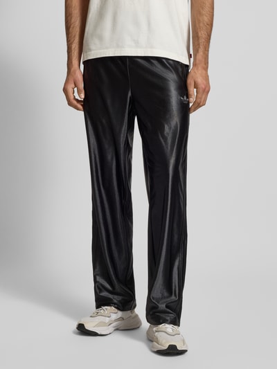 Męski model nosi czarne spodnie treningowe adidas Originals Trainingshose mit Logo Modell 'FIREBIRD', krój regular fit, logo, elastyczny pas, długość wewnętrzna nogawki 79 cm dla rozmiaru M; zdjęcie: detal z przodu, widok od pasa w dół.